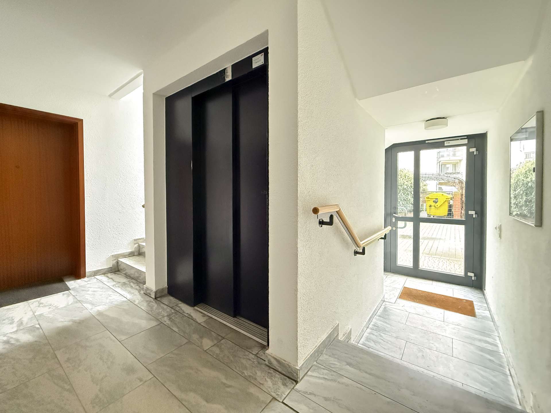 Lobby hallway with a closed black elevator and a staircase nearby, marble tile floor, and a glass exit door at the end of the hall.
