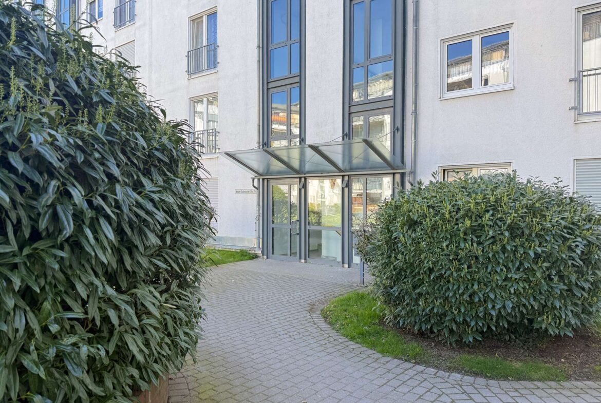 Entrance to a modern apartment building with a glass canopy over the doors and tall windows reflecting the sky, flanked by dense green shrubs on a paved walkway.