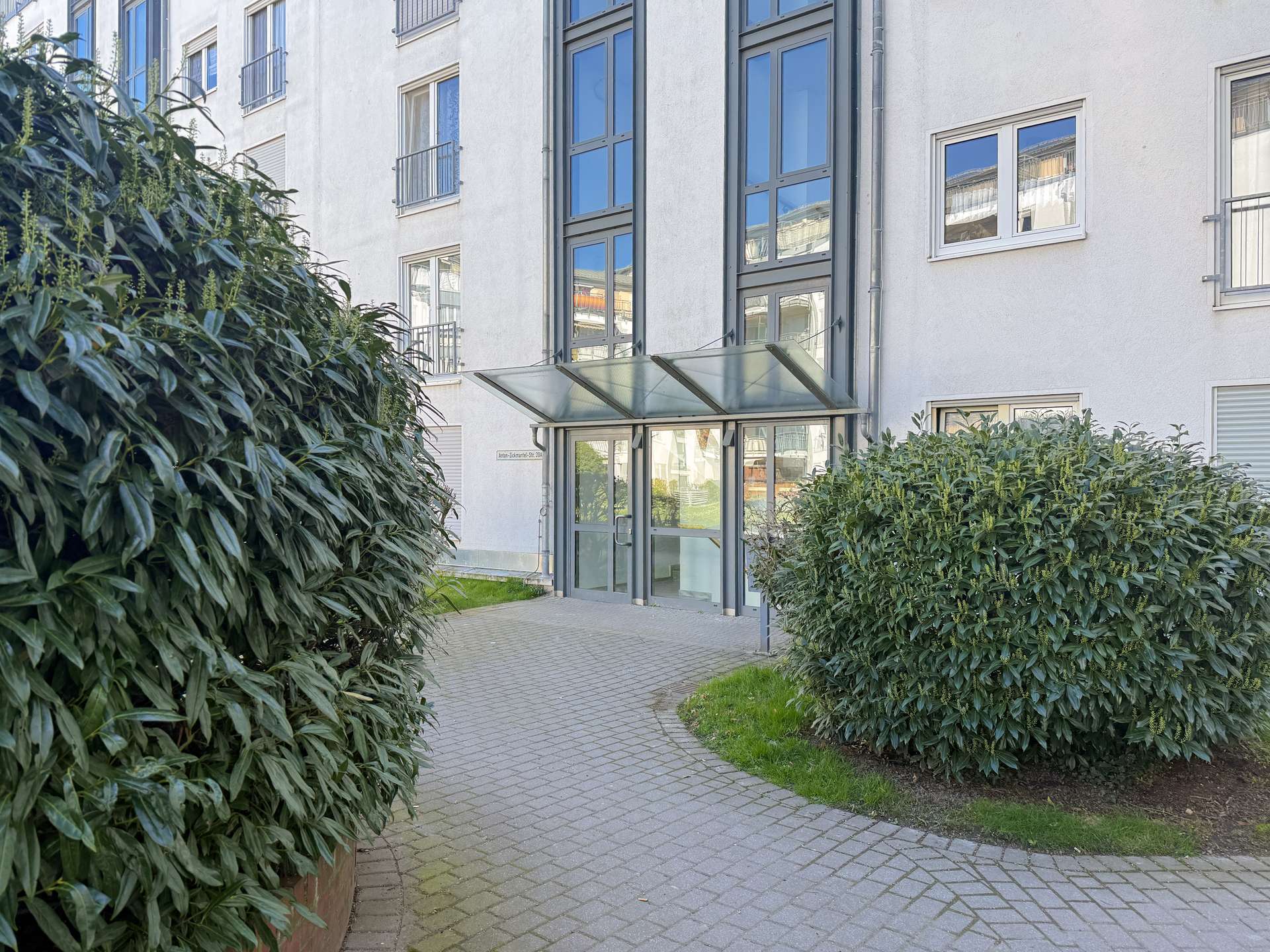 Entrance to a modern apartment building with a glass canopy over the doors and tall windows reflecting the sky, flanked by dense green shrubs on a paved walkway.
