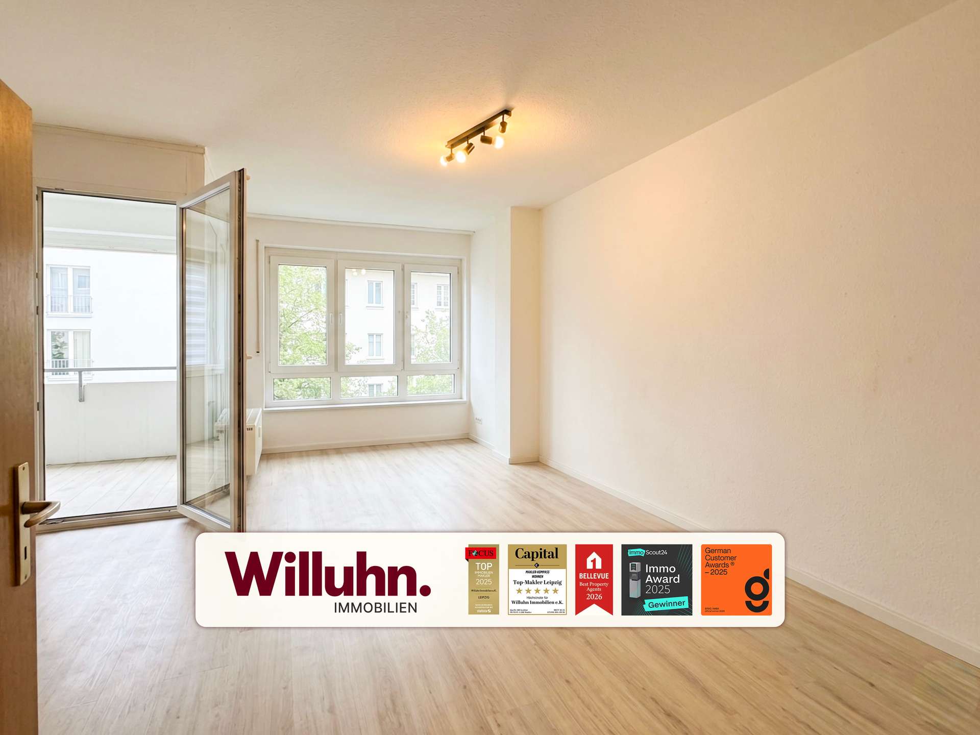 Bright, empty living room with large windows and an open glass balcony door; Real estate banner reads 'Willuhn Immobilien' with award badges along the bottom edge.