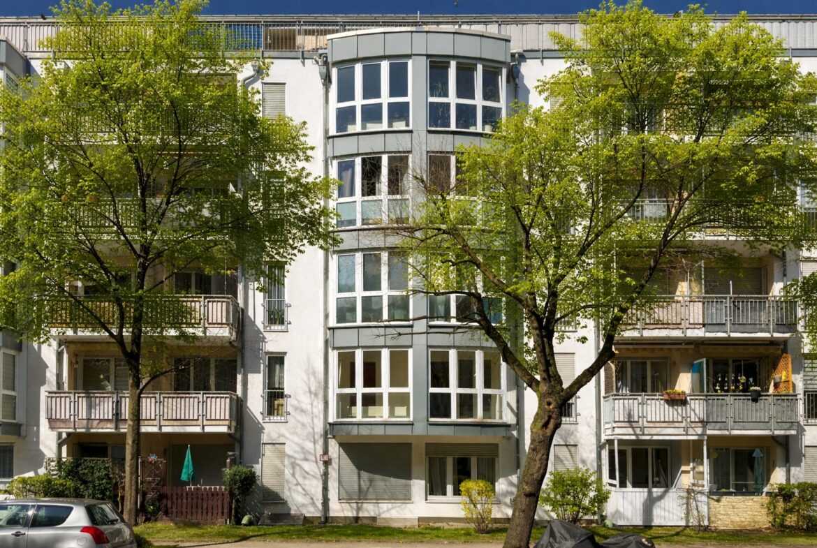 Facade of a white apartment building with large bay windows, framed by green trees in front.