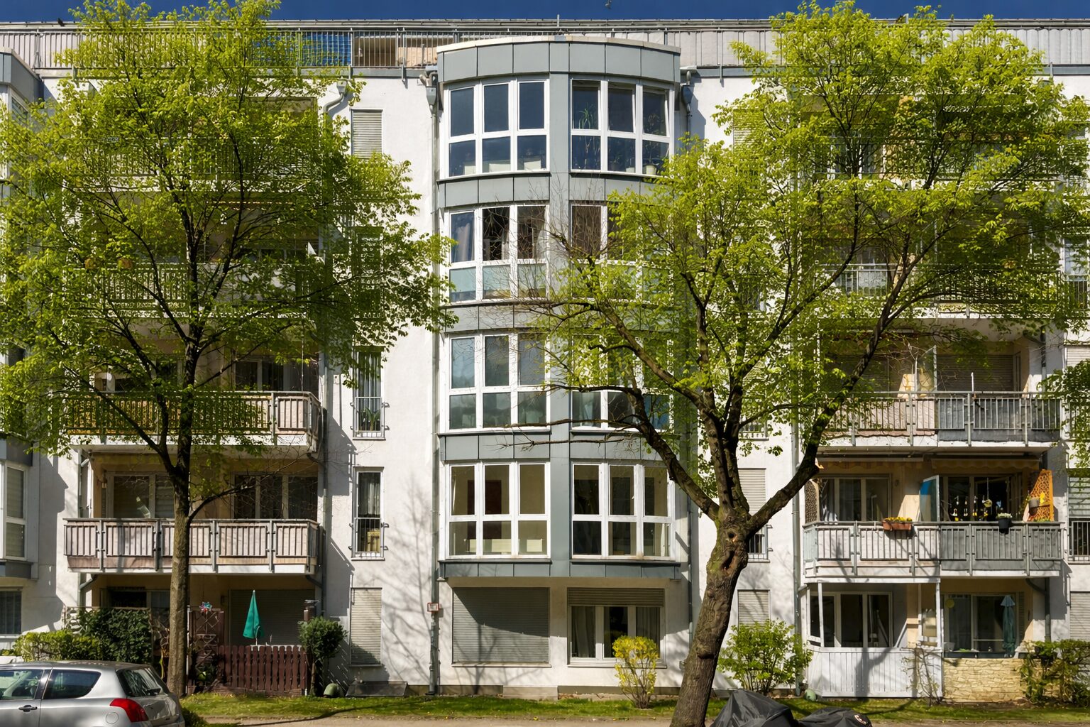 Facade of a white apartment building with large bay windows, framed by green trees in front.