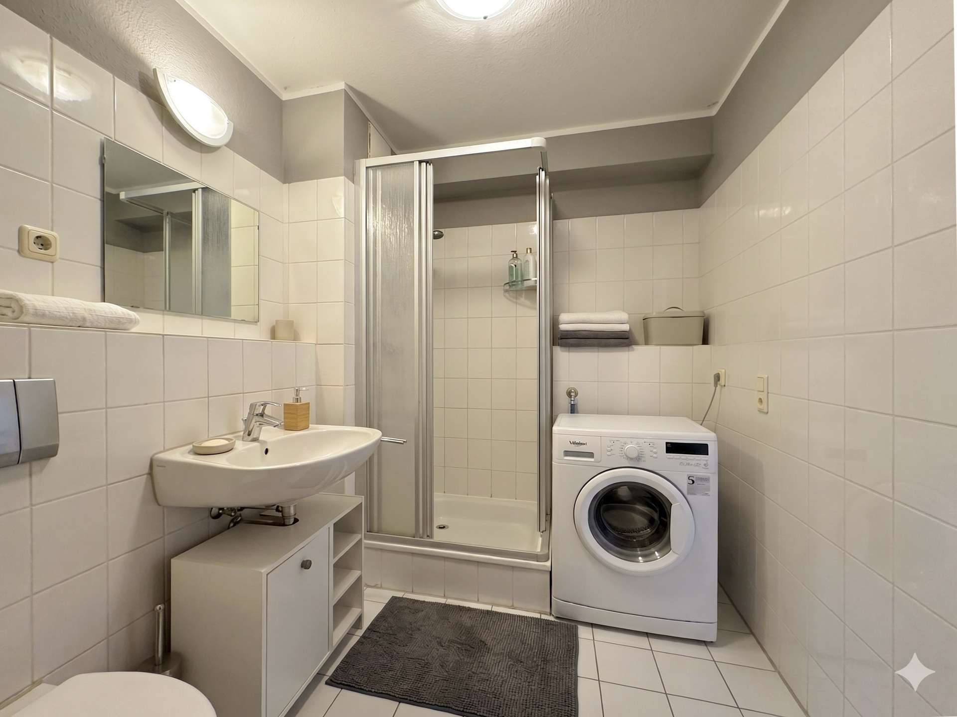 Compact bathroom with a washing machine beside a glass shower enclosure and white tiled walls; grey bath mats on the floor.