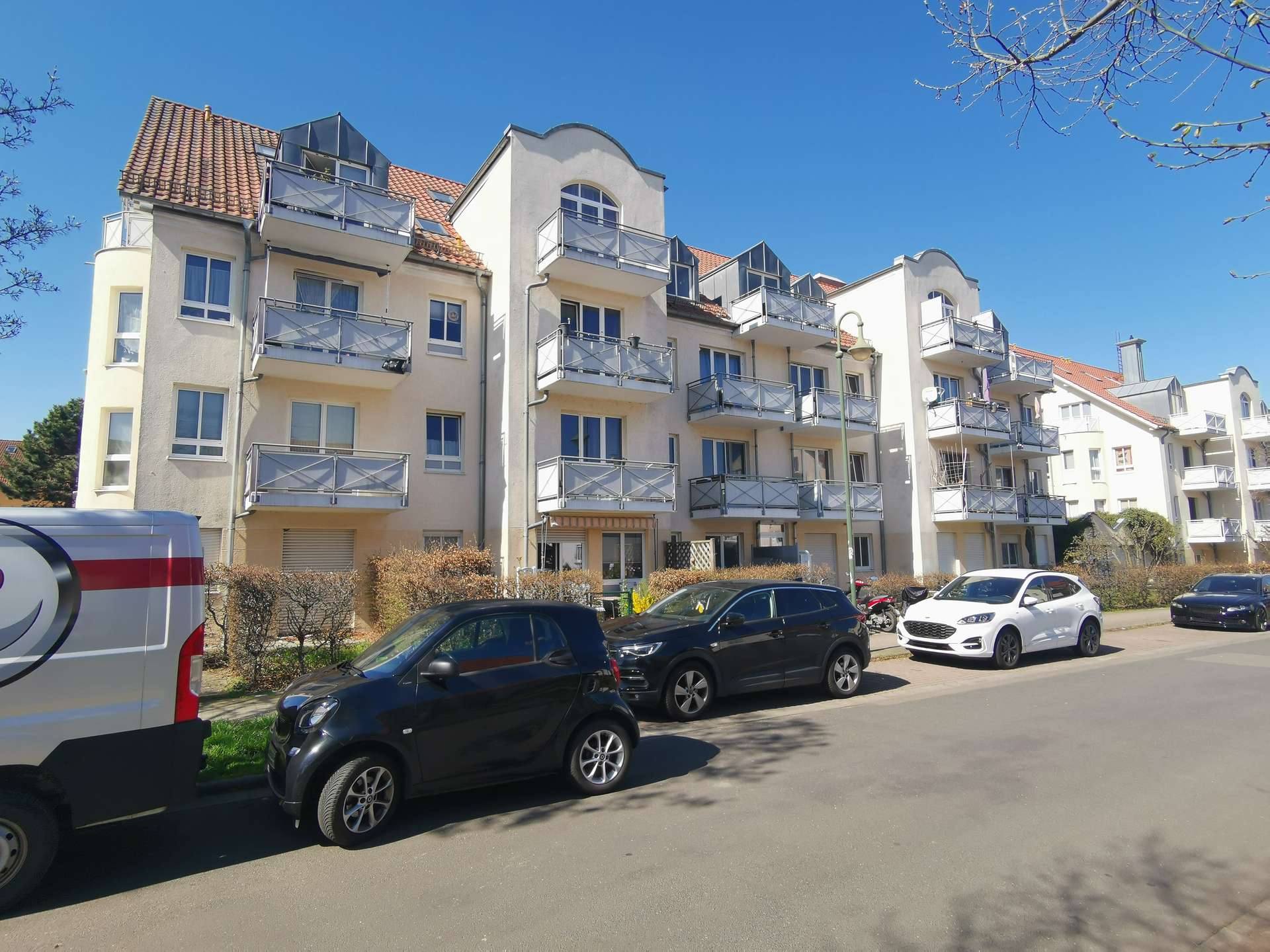 Apartment building with multiple balconies and a blue sky, parked cars lining the street in front of beige facades.