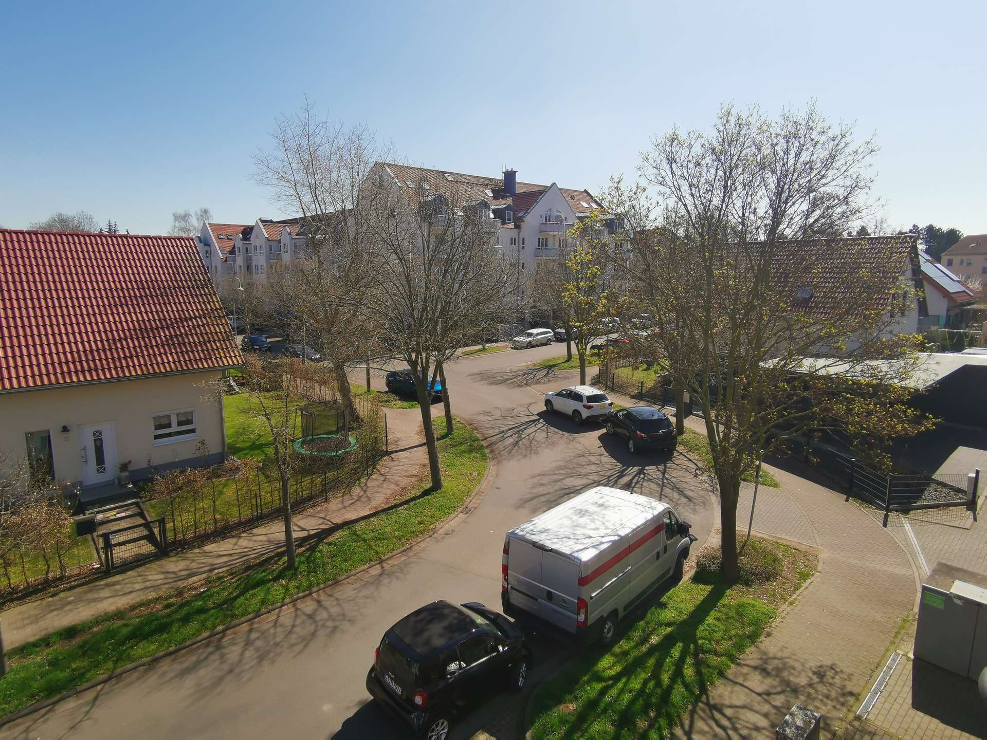 Aerial view of a residential street with red-tiled roofs, leafless trees, and parked cars on both sides under a clear blue sky.