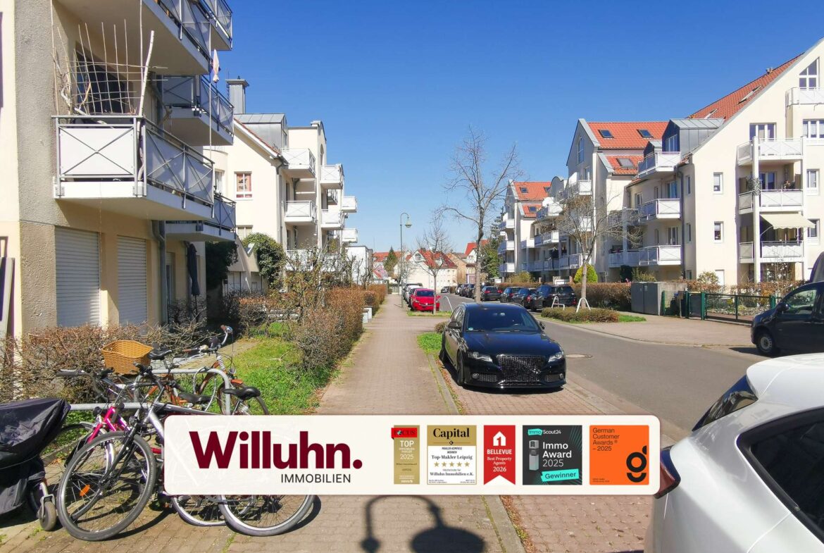 Residential street with modern beige apartment buildings on both sides, balconies visible, blue sky, and bikes parked along the foreground. A large Willuhn Immobilien banner spans the bottom of the image.
