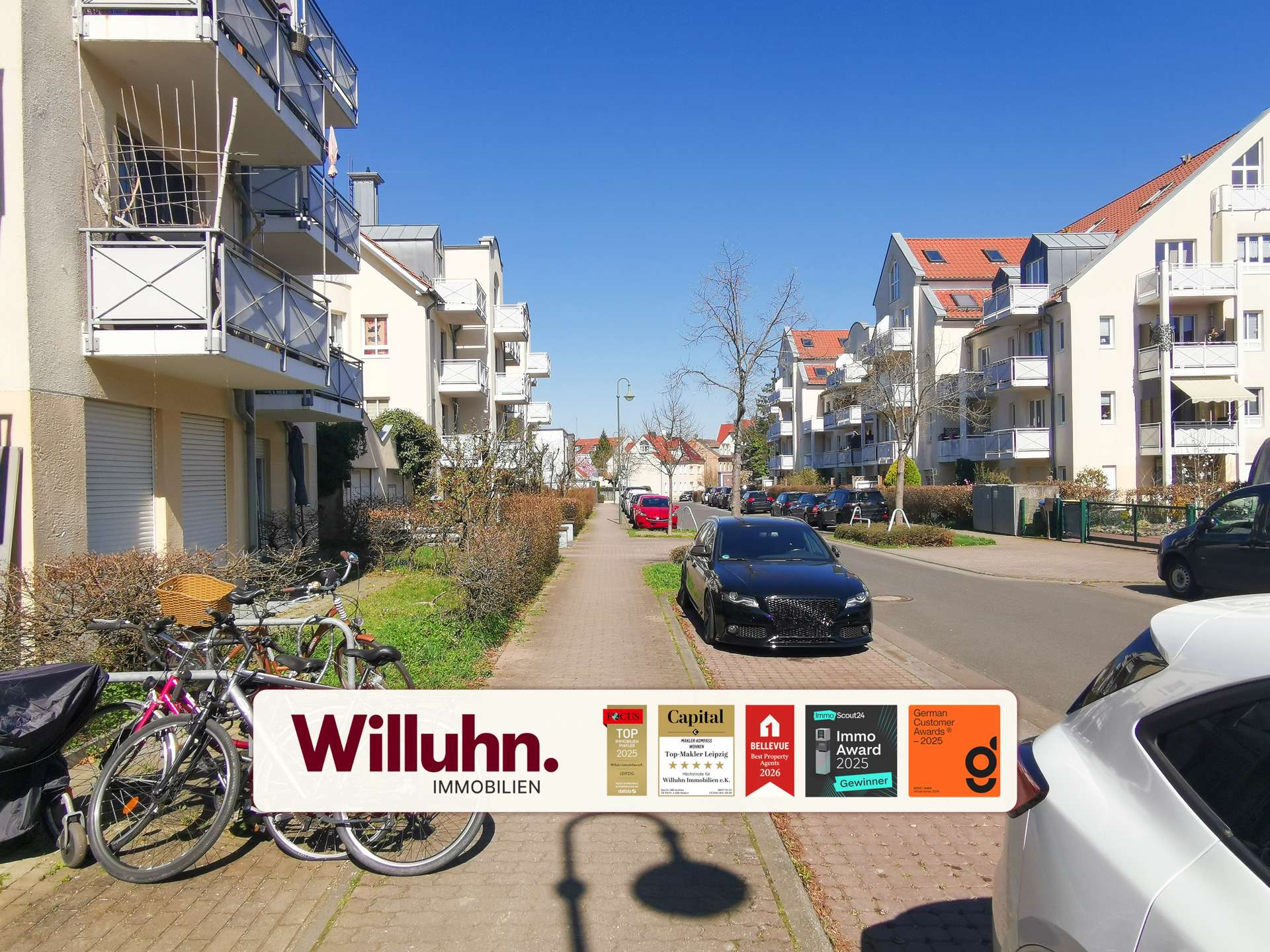Residential street with modern beige apartment buildings on both sides, balconies visible, blue sky, and bikes parked along the foreground. A large Willuhn Immobilien banner spans the bottom of the image.