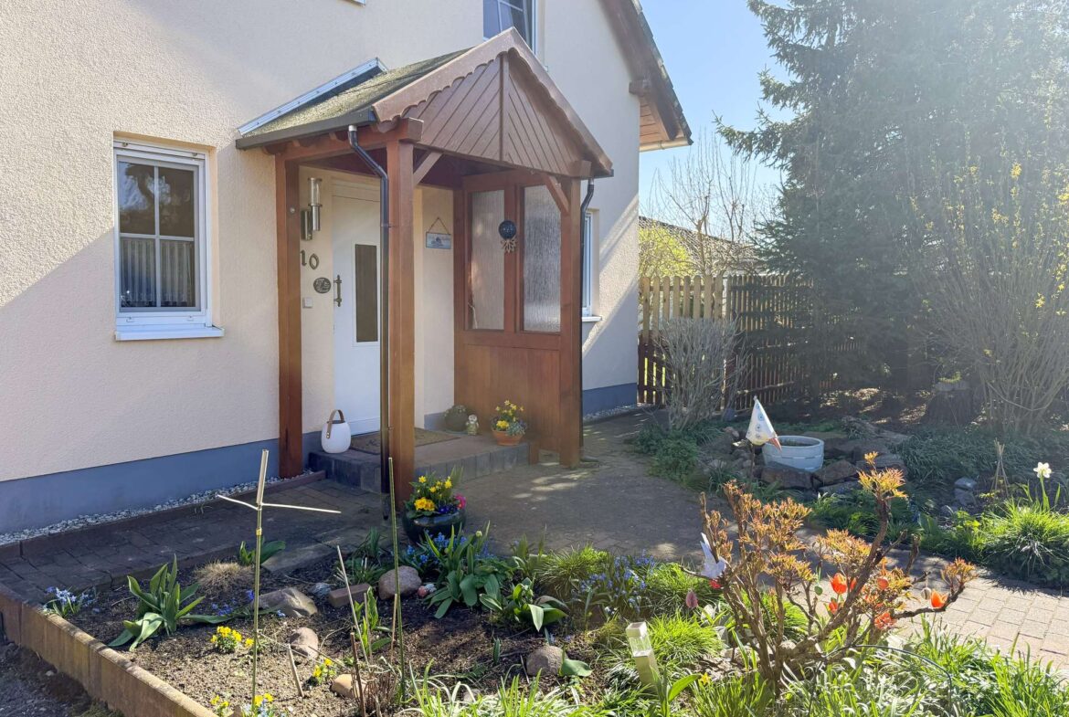 House front entrance with a wooden porch, beige exterior walls, and a small garden bed full of daffodils and other flowers in the foreground.