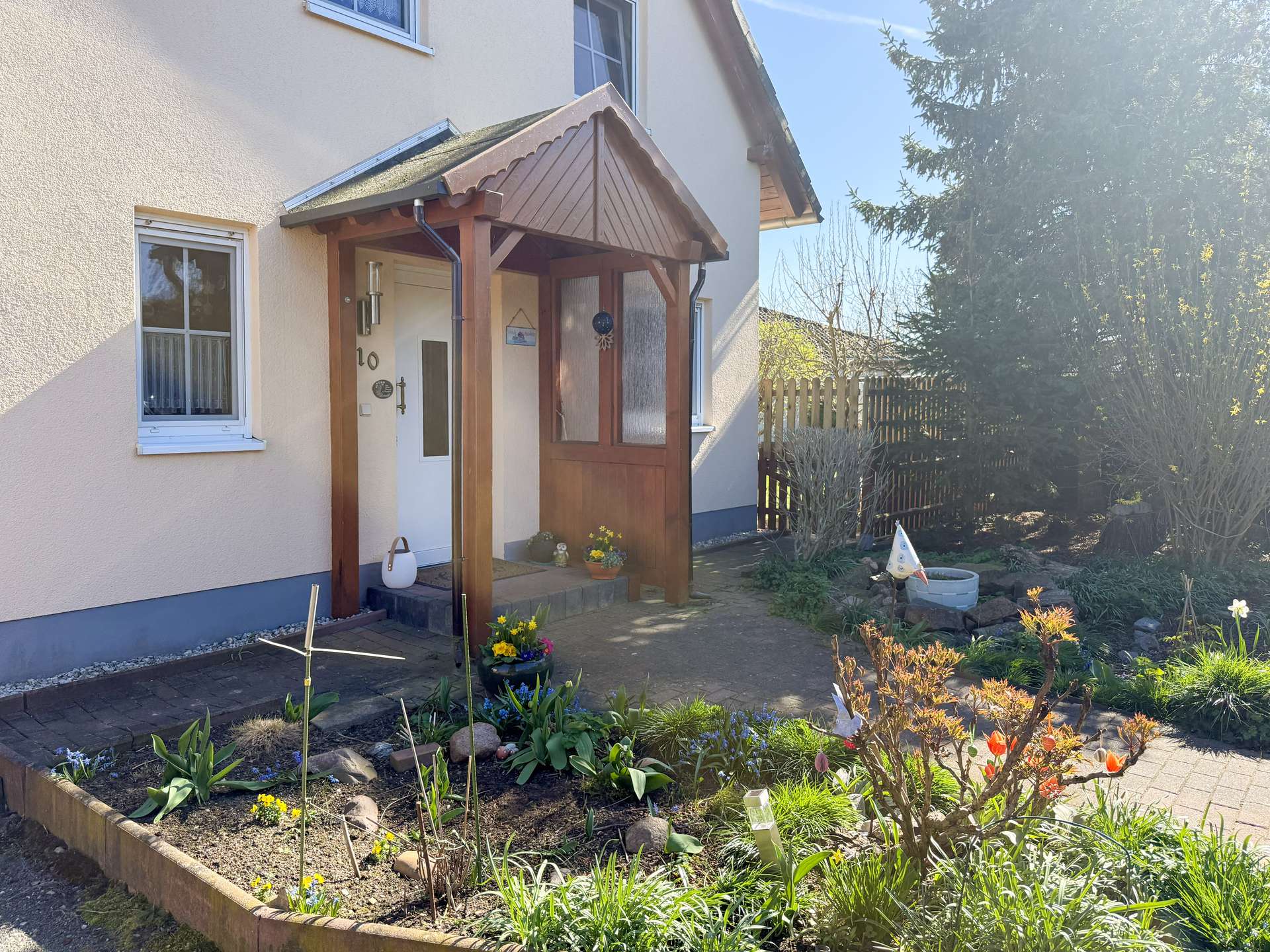 House front entrance with a wooden porch, beige exterior walls, and a small garden bed full of daffodils and other flowers in the foreground.