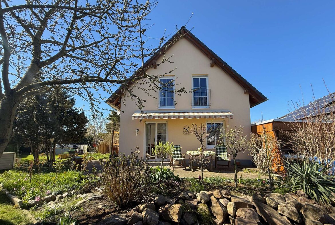 Beige two-story house with a striped awning over a door, a small balcony, and a garden with rocks and blooming plants in the foreground.