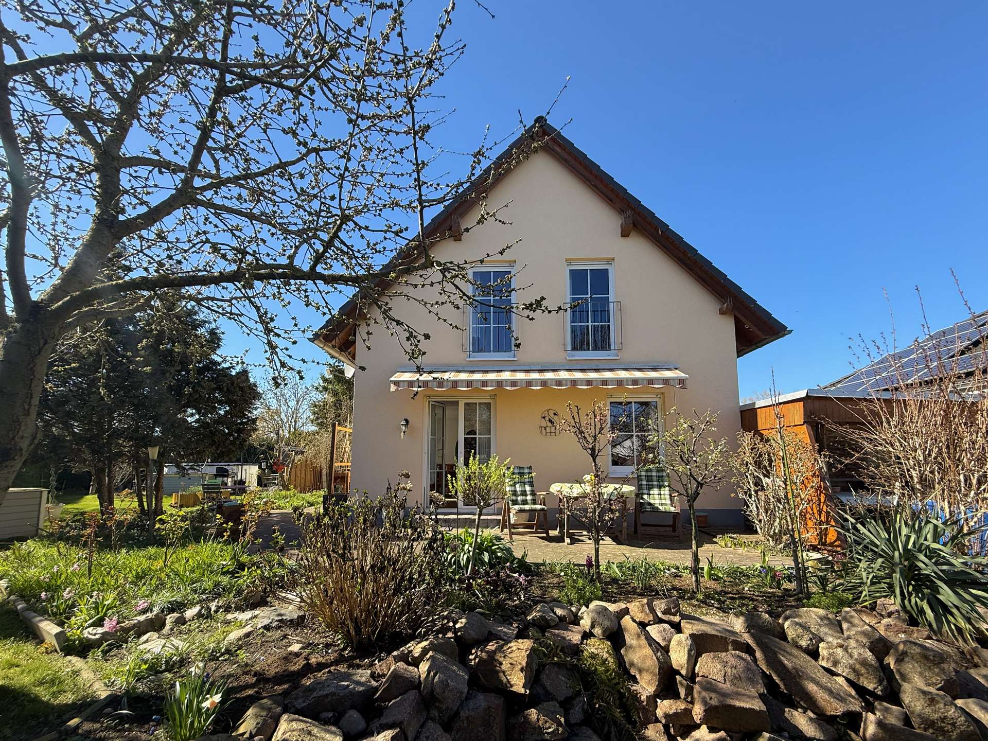 Beige two-story house with a striped awning over a door, a small balcony, and a garden with rocks and blooming plants in the foreground.