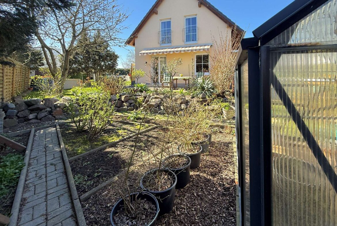 Front yard view of a two-story beige house with a striped awning over a glass patio door and potted trees along a stone-bordered garden path on a sunny day