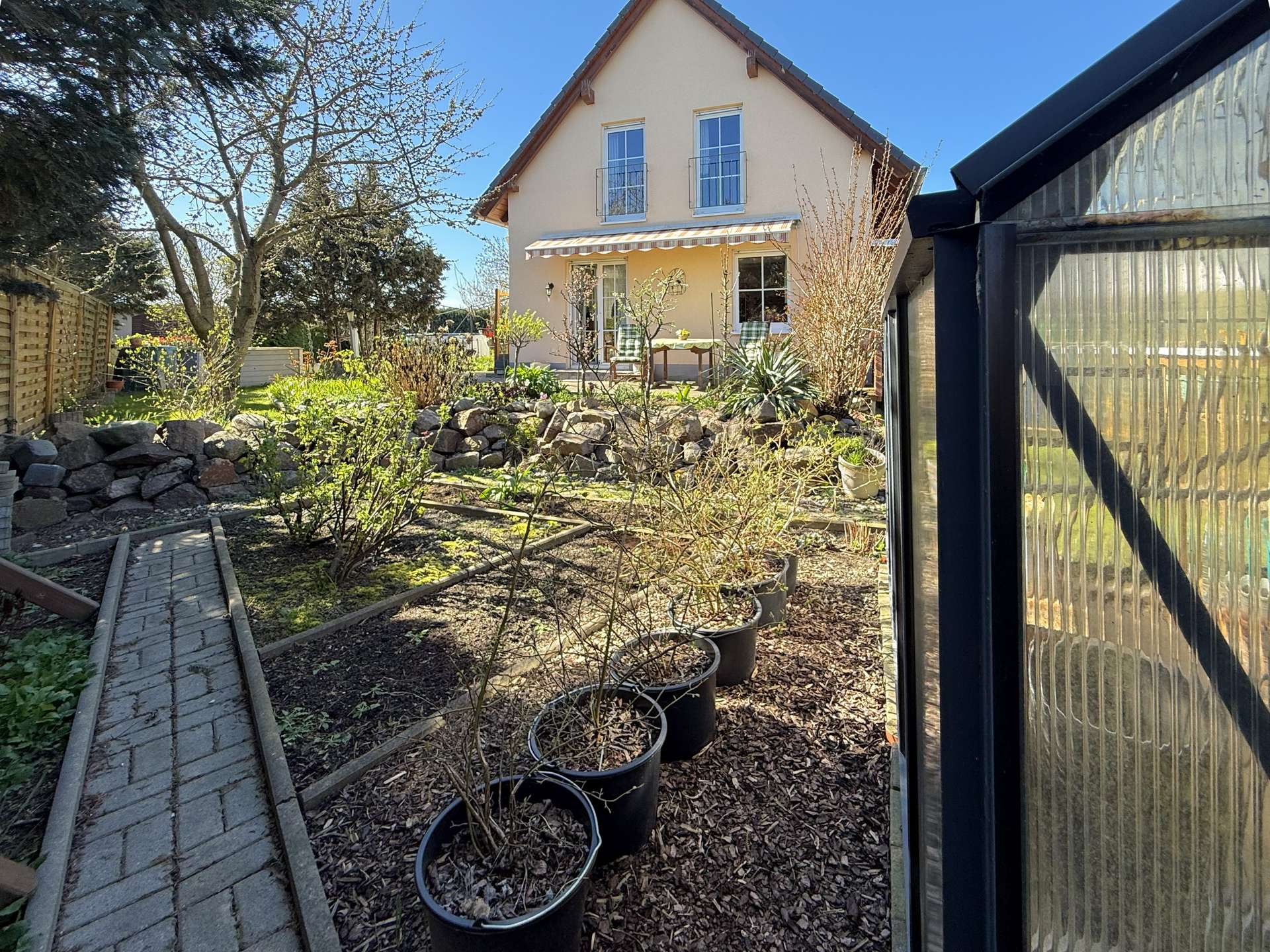 Front yard view of a two-story beige house with a striped awning over a glass patio door and potted trees along a stone-bordered garden path on a sunny day