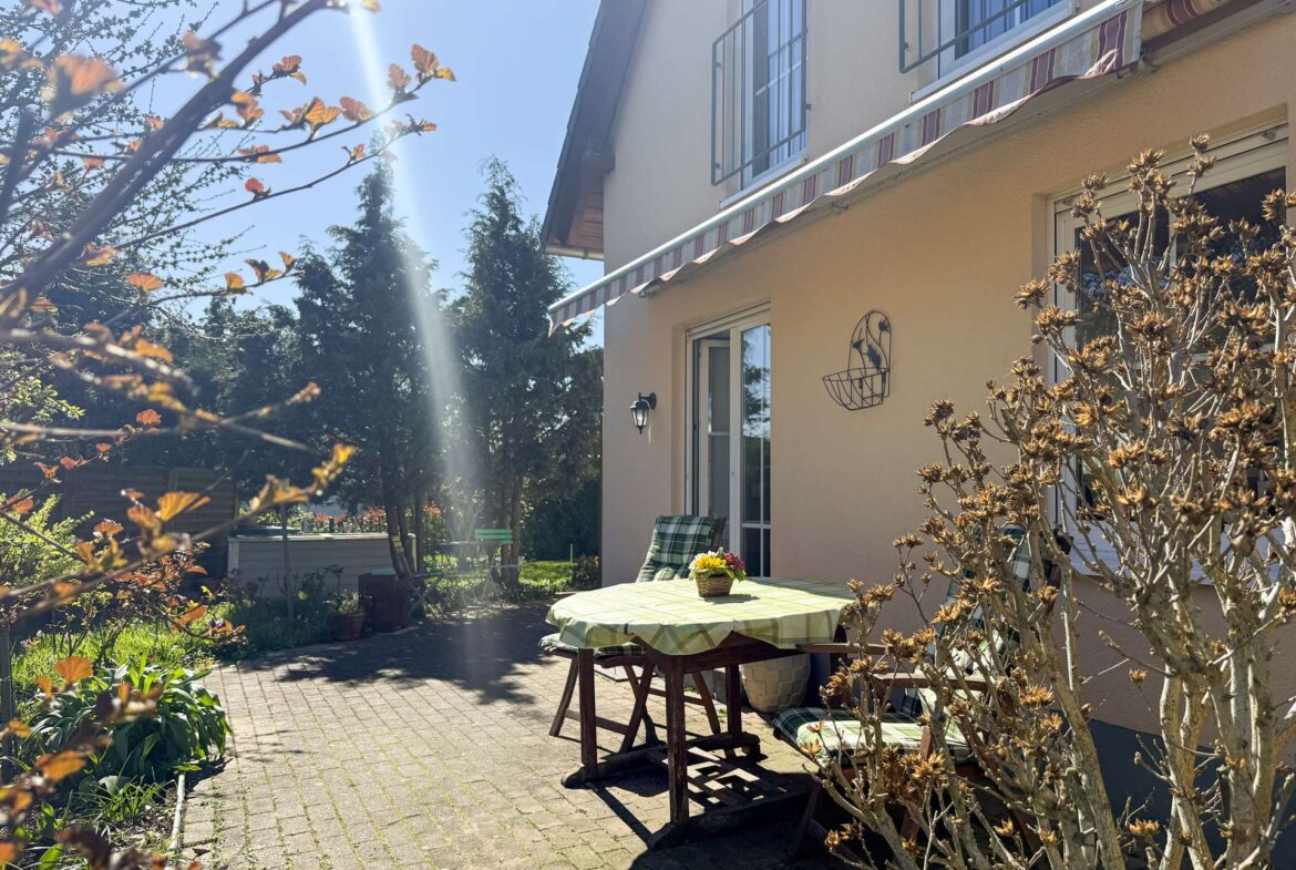 Sunny backyard patio with a round table covered by a green checkered tablecloth, a small plant basket in the center, and beige house walls with windows and a striped awning above. A leafless shrub in the foreground and seating area in the background.