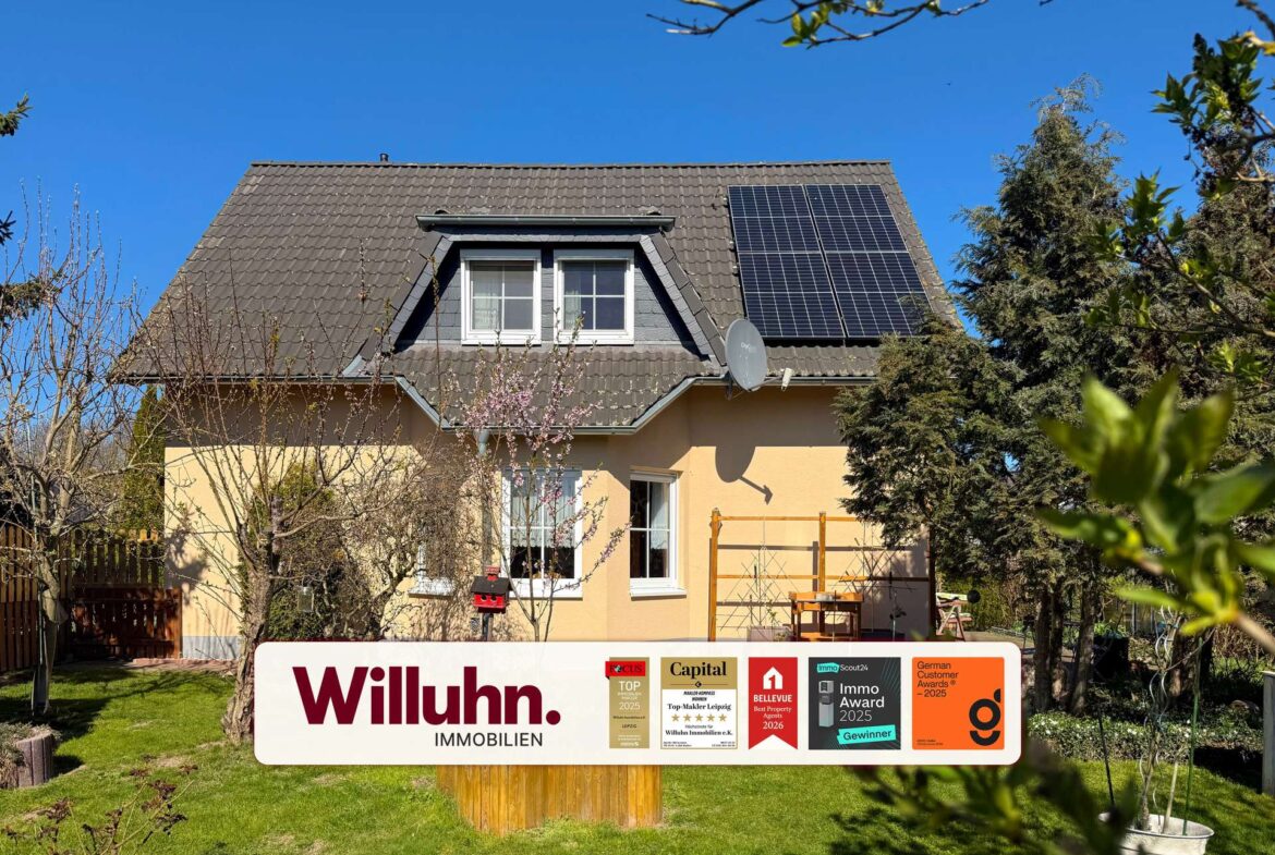Two-story house with a brown tiled roof, dormer window, and solar panels under a bright blue sky; a large Willuhn Immobilien sign in the front yard.