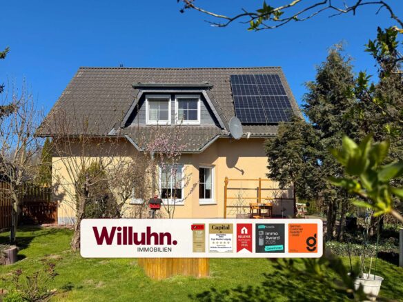Two-story house with a brown tiled roof, dormer window, and solar panels under a bright blue sky; a large Willuhn Immobilien sign in the front yard.