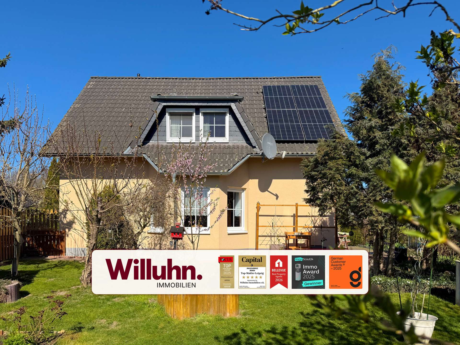 Two-story house with a brown tiled roof, dormer window, and solar panels under a bright blue sky; a large Willuhn Immobilien sign in the front yard.