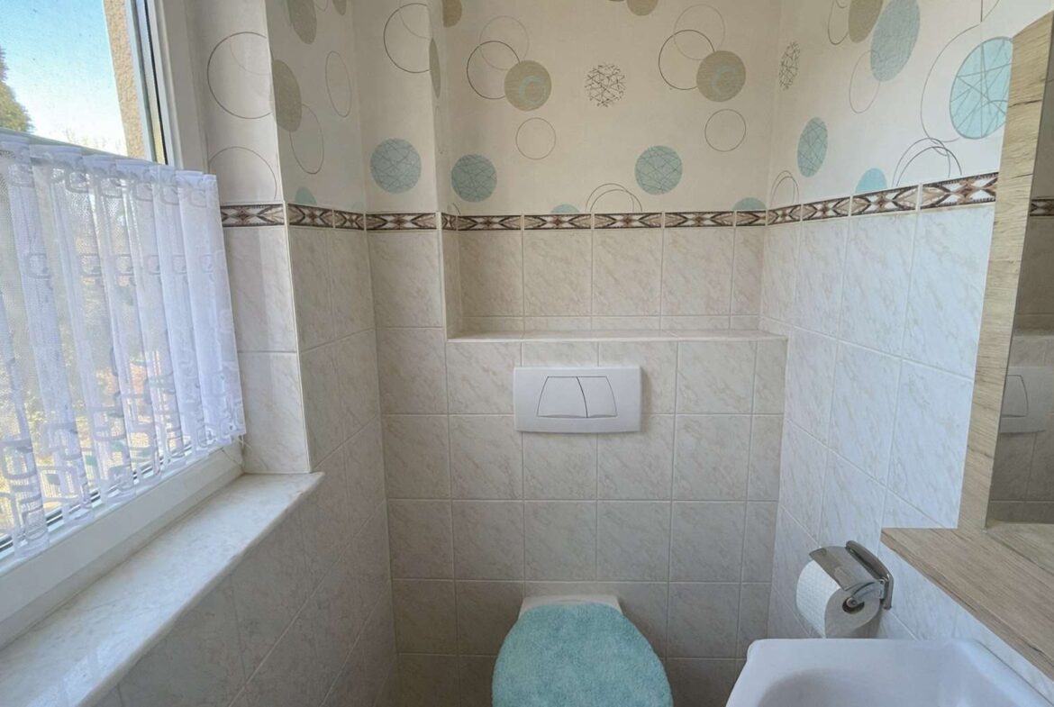 Small bathroom with wall-mounted toilet and turquoise toilet seat cover; window with lace curtain on left; tiled walls and decorative wallpaper with circles above the tiles.