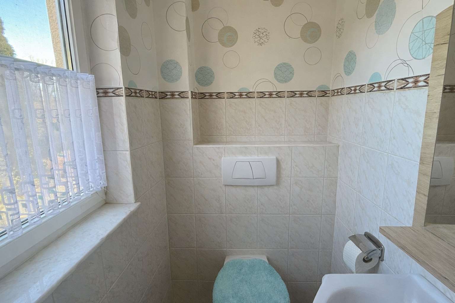 Small bathroom with wall-mounted toilet and turquoise toilet seat cover; window with lace curtain on left; tiled walls and decorative wallpaper with circles above the tiles.