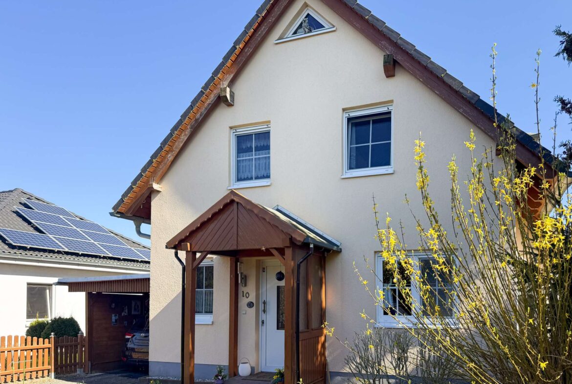 Front view of a beige two-story house with a steep gabled roof, wooden porch, and flowering bushes in the yard.