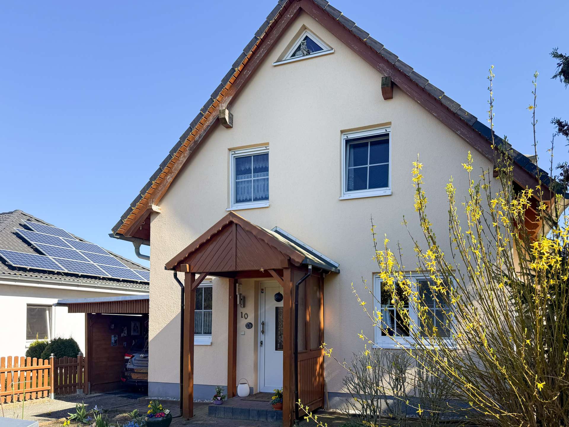 Front view of a beige two-story house with a steep gabled roof, wooden porch, and flowering bushes in the yard.