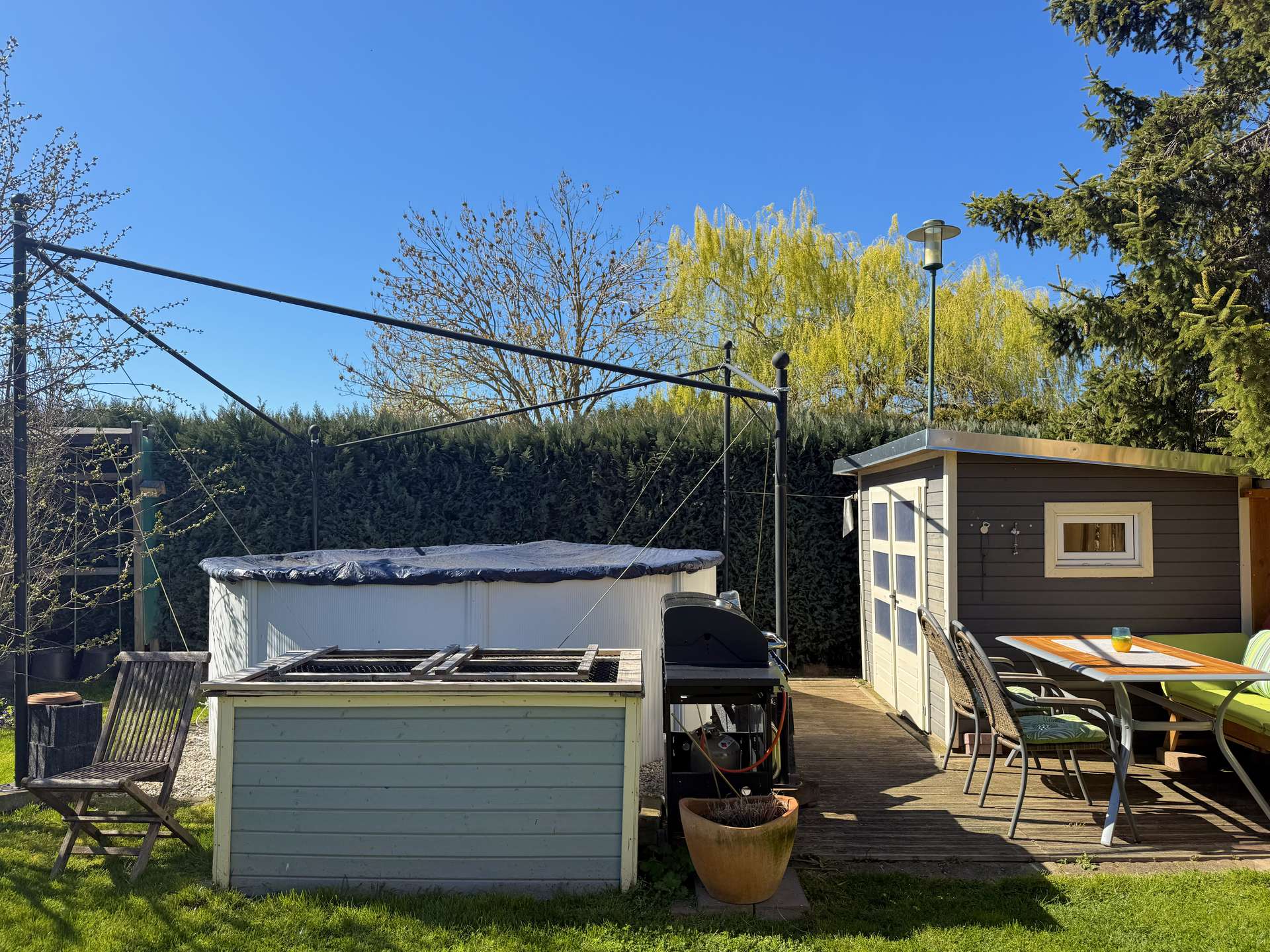 Backyard scene with a small shed, patio table and chairs, a covered pool, and potted plant under a clear blue sky.