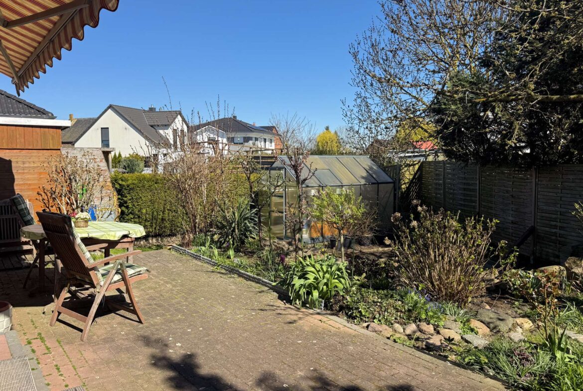Sunny backyard patio with wooden chairs around a table, garden beds and a greenhouse in the background.