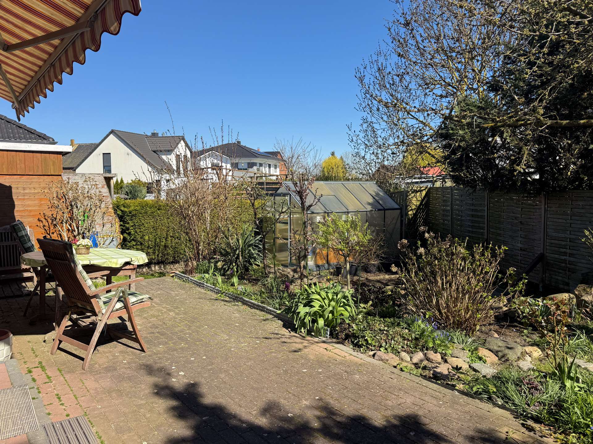 Sunny backyard patio with wooden chairs around a table, garden beds and a greenhouse in the background.