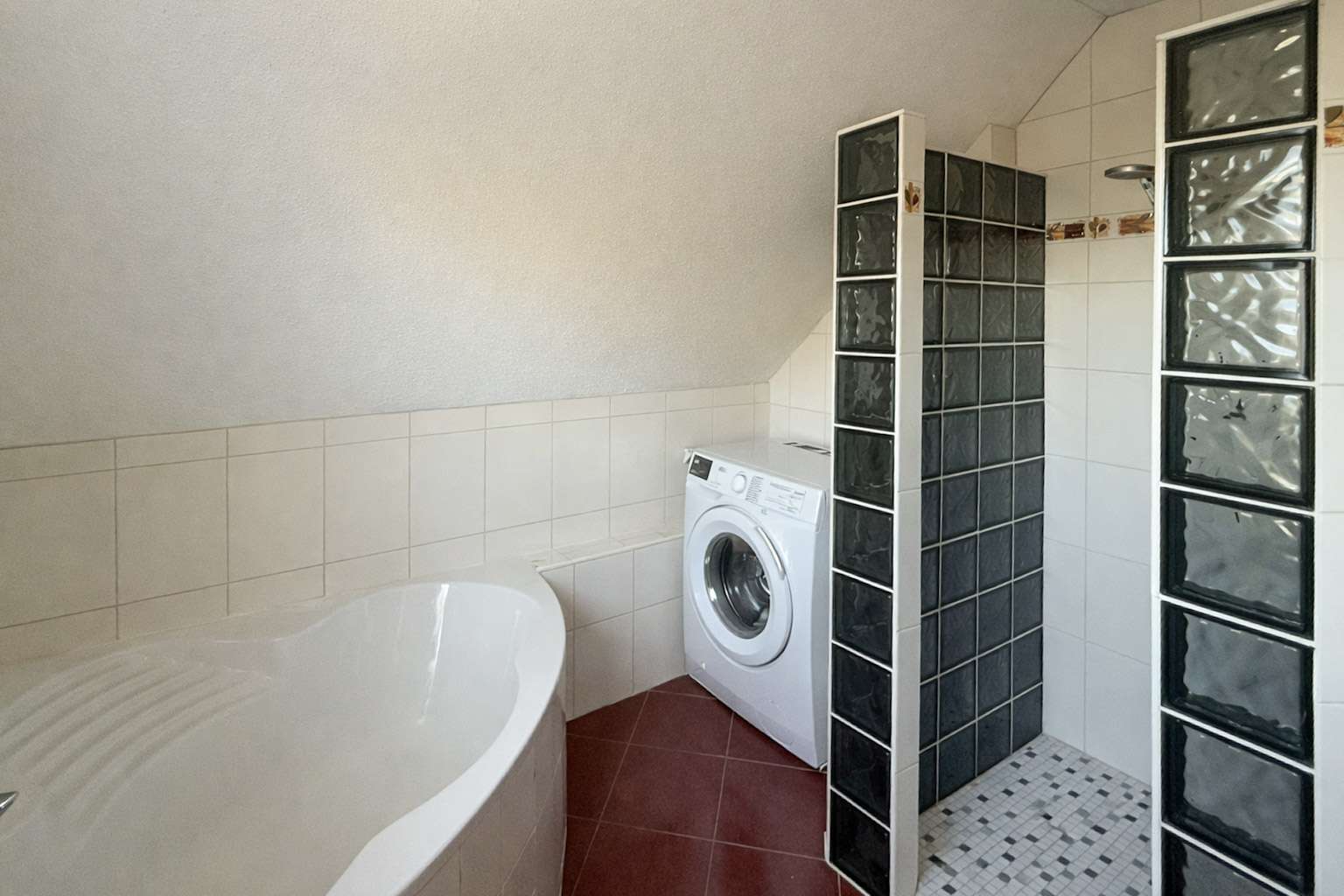 Bathroom corner with a front-loading washing machine next to a curved white bathtub and glass block partitions; red floor tiles.
