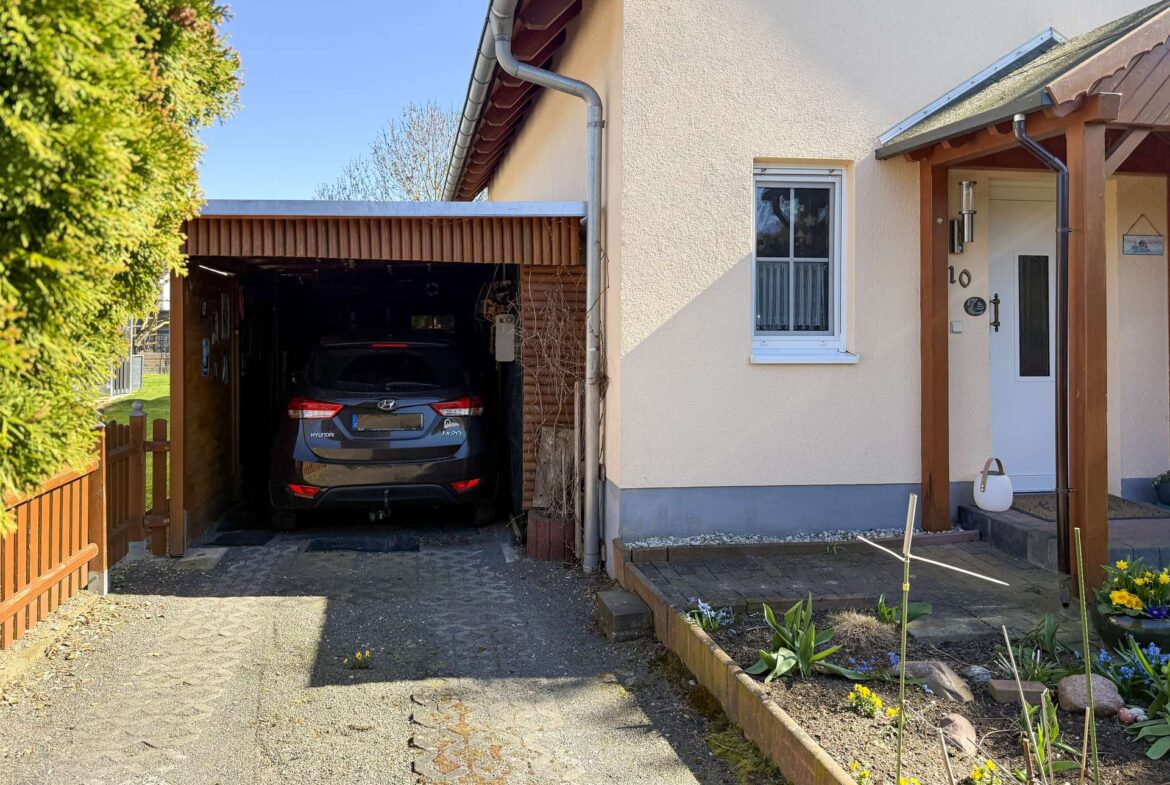 A blue Hyundai SUV parked in a shaded carport beside a beige house with a small garden bed along the walkway to the front door.