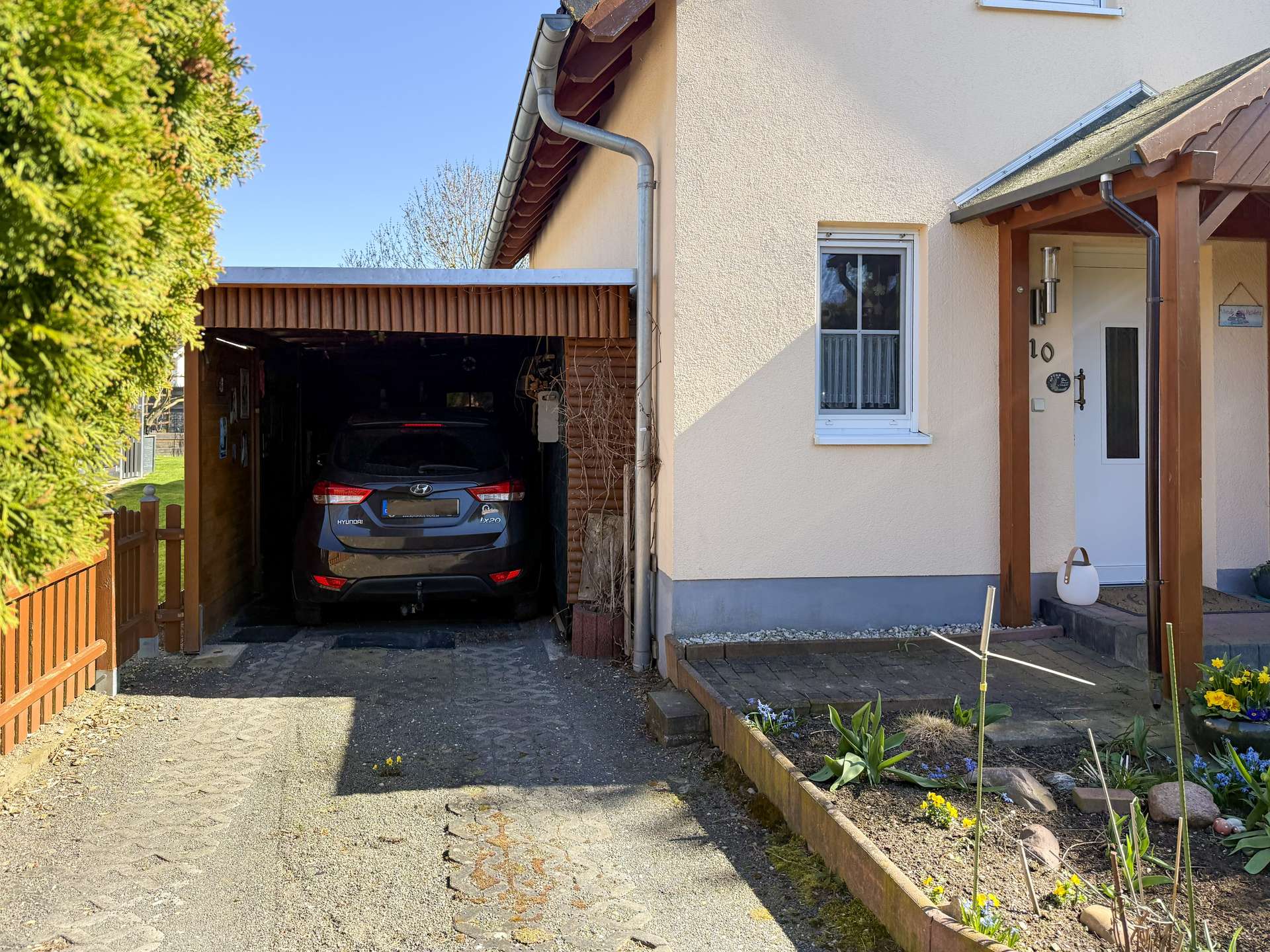 A blue Hyundai SUV parked in a shaded carport beside a beige house with a small garden bed along the walkway to the front door.