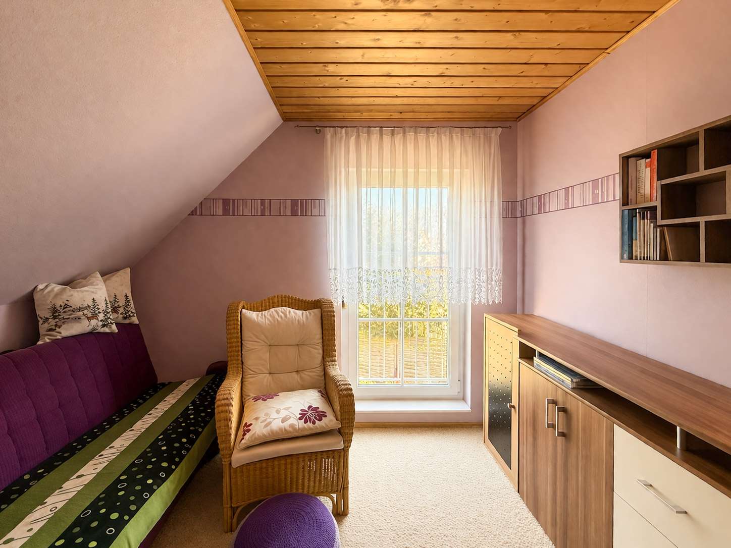 Cozy attic room with purple walls, wooden sloped ceiling, and a window with sheer lace curtains; wicker chair and purple sofa with cushions nearby.