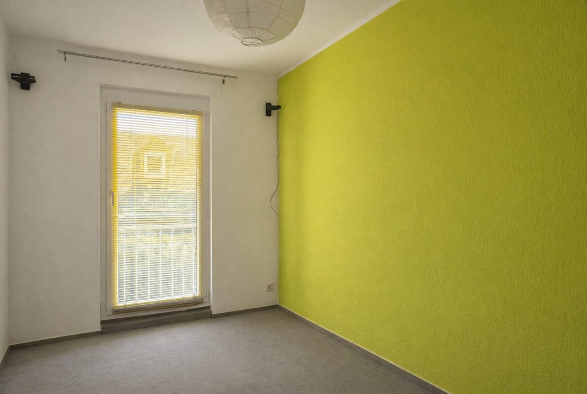 Small empty room with a bright lime green accent wall on the right, white walls, gray carpet, and a window with yellow blinds. A round ceiling light hangs above.