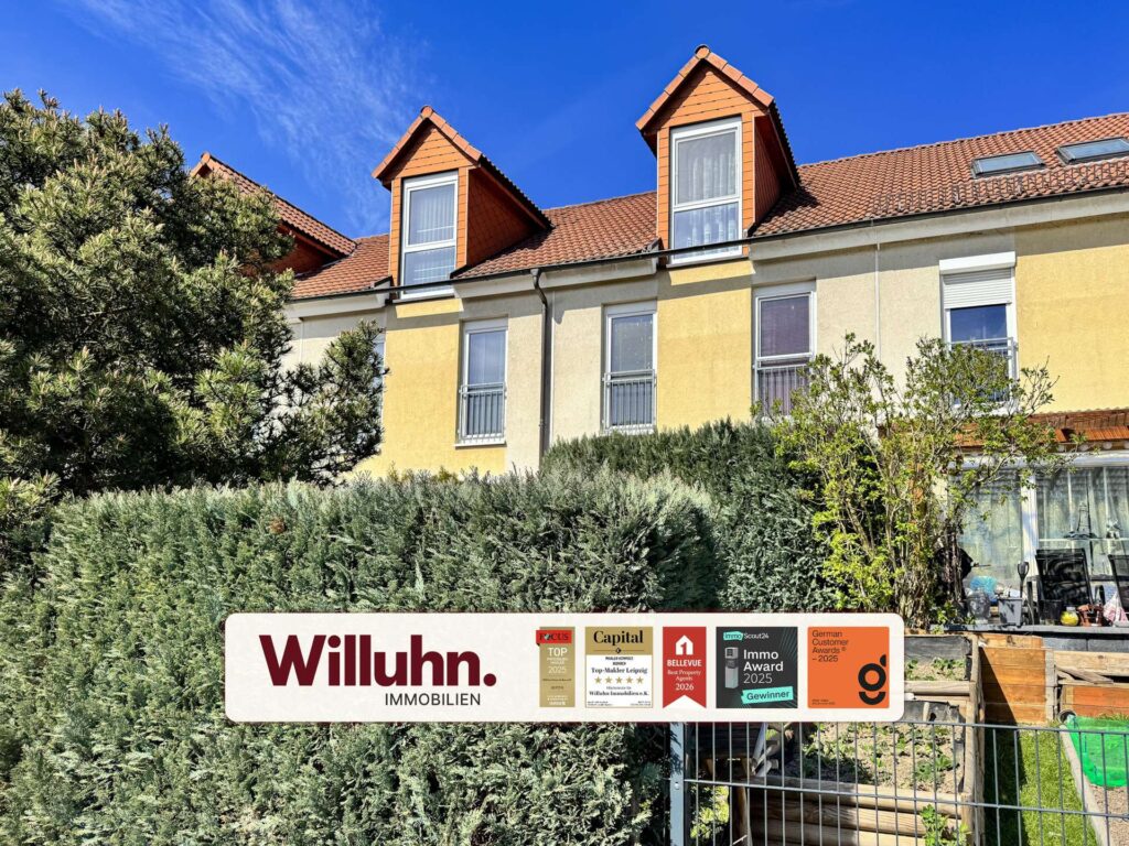 Facade of a yellow apartment building with red tile roof and dormer windows, set against a blue sky, with a Willuhn Immobilien sign in front.
