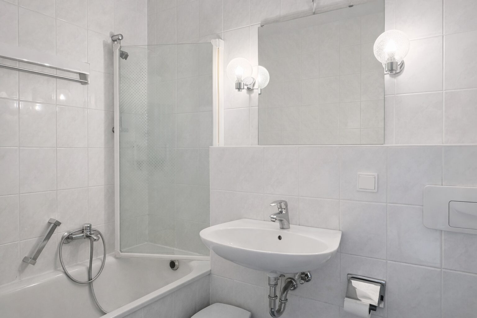 Bright white bathroom featuring a bathtub with a glass shower screen, wall mirror, and pedestal sink with exposed chrome plumbing.
