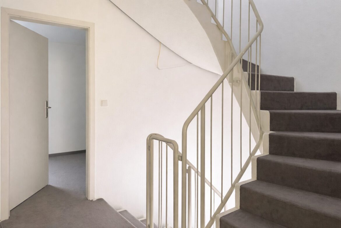 Interior stairwell with beige walls, a carpeted staircase on the right, and an open door to a small room on the left; a railing runs along the stairs.