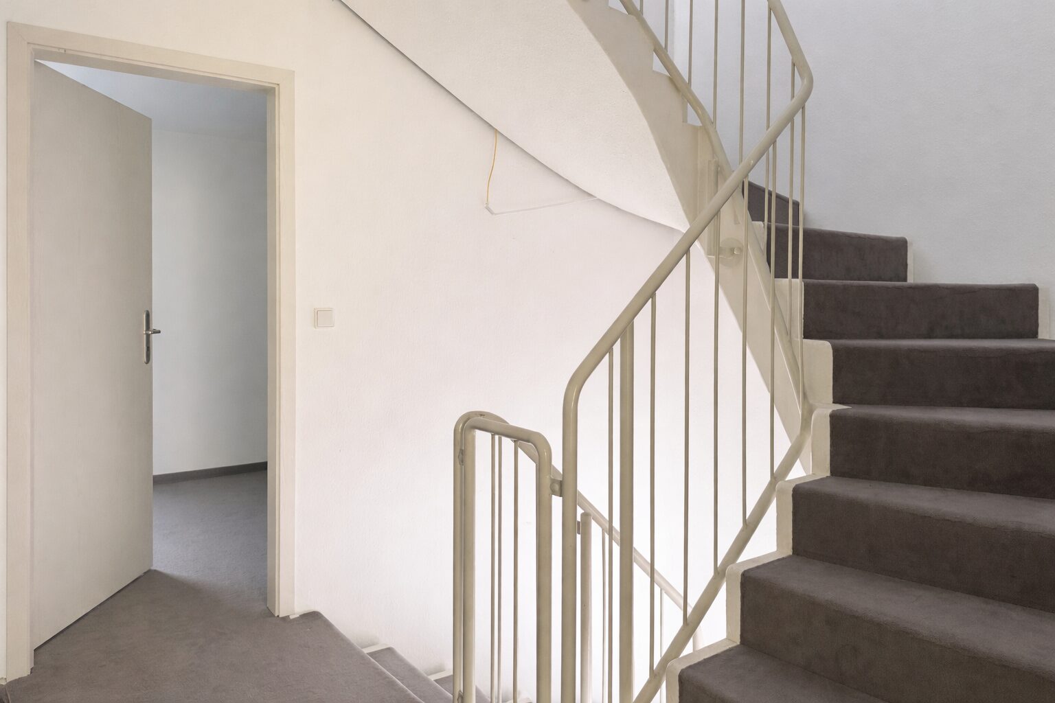 Interior stairwell with beige walls, a carpeted staircase on the right, and an open door to a small room on the left; a railing runs along the stairs.