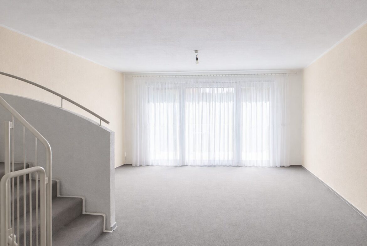 Empty living room with grey carpet, cream walls, and a curved white staircase on the left; large window with sheer white curtains glowing with daylight.