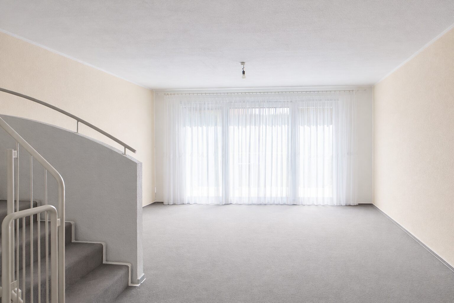 Empty living room with grey carpet, cream walls, and a curved white staircase on the left; large window with sheer white curtains glowing with daylight.
