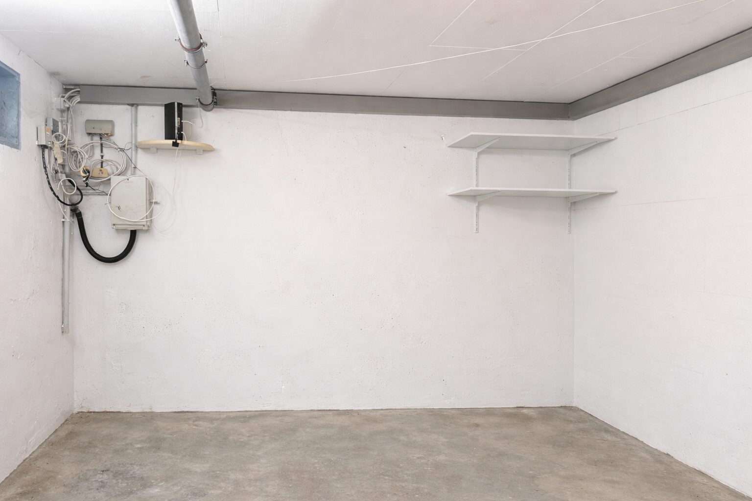 Bare basement utility room with exposed wiring and a vertical electrical panel on the left wall, white walls, and two empty white wall shelves on the right.