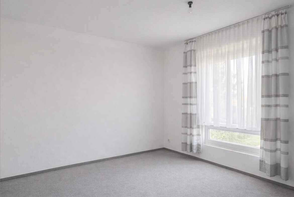 Empty white room with a large window dressed in sheer white curtains and gray-striped drapes, grey carpeted floor.