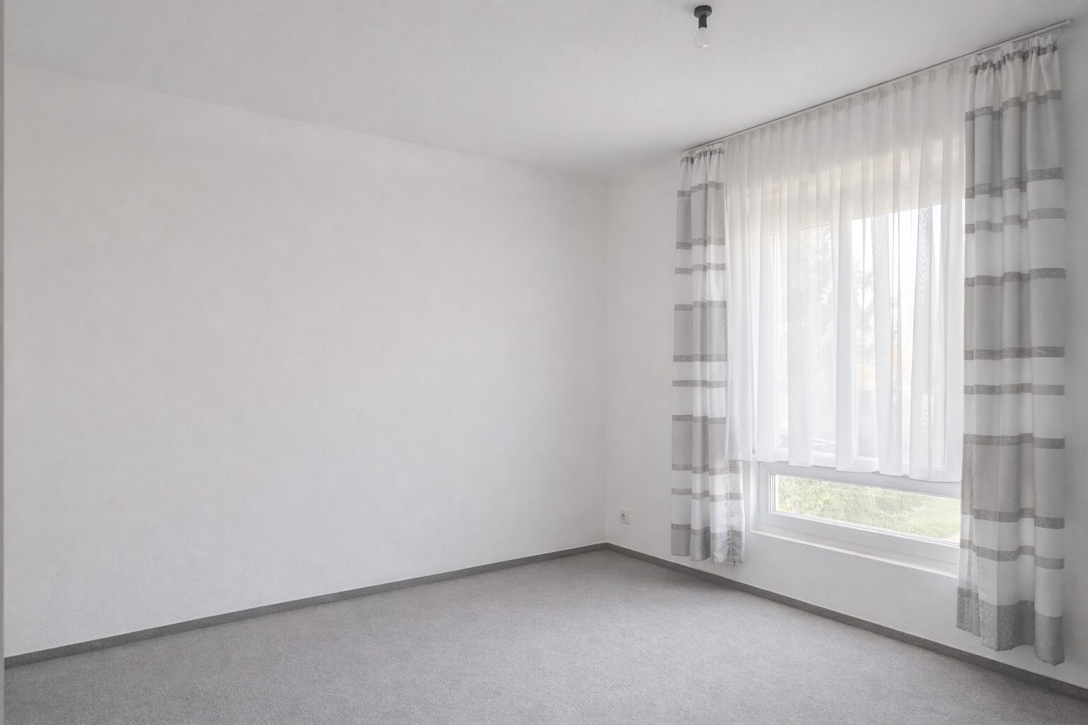 Empty white room with a large window dressed in sheer white curtains and gray-striped drapes, grey carpeted floor.