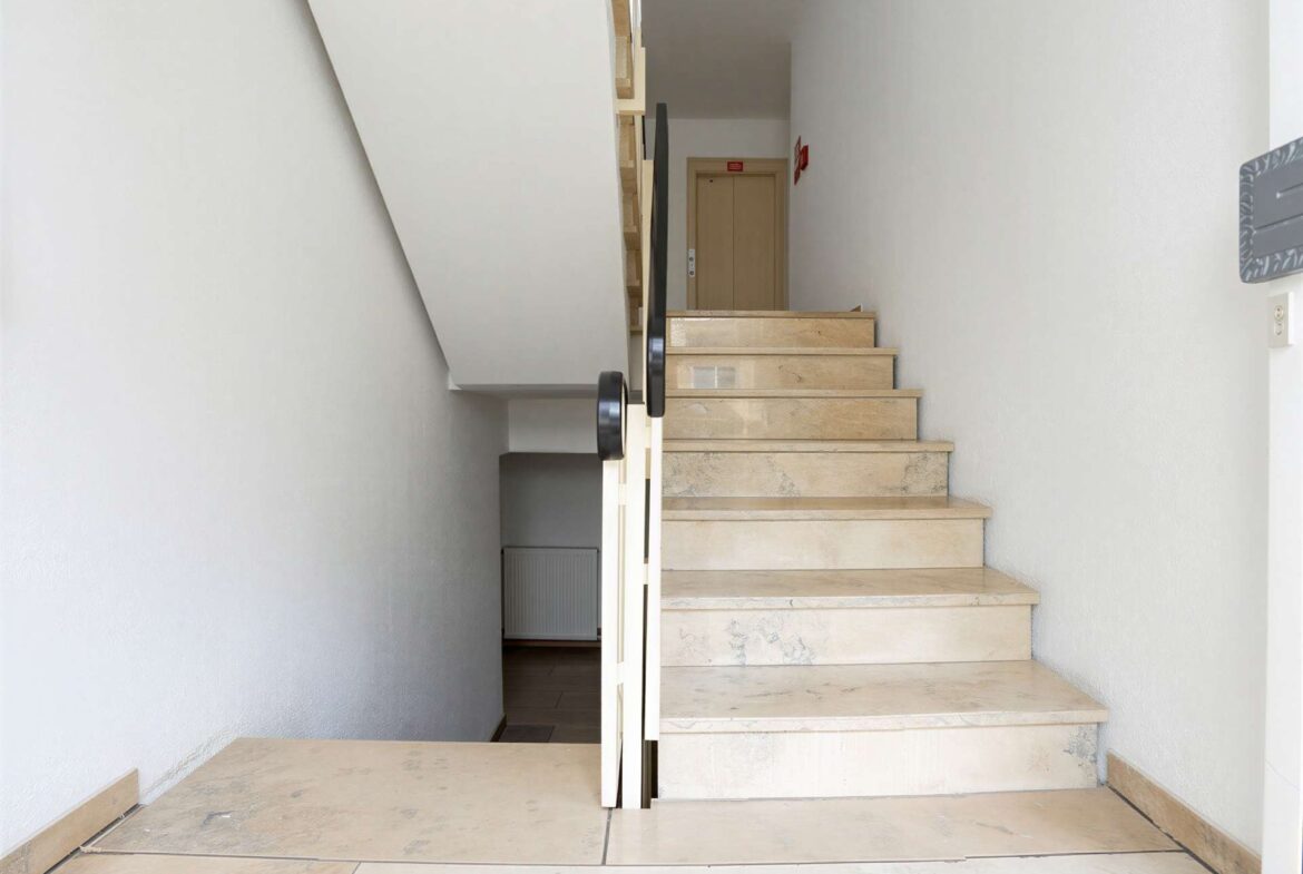 Beige marble staircase in a narrow white-walled hallway, with a black handrail leading up to a wooden door at the top.