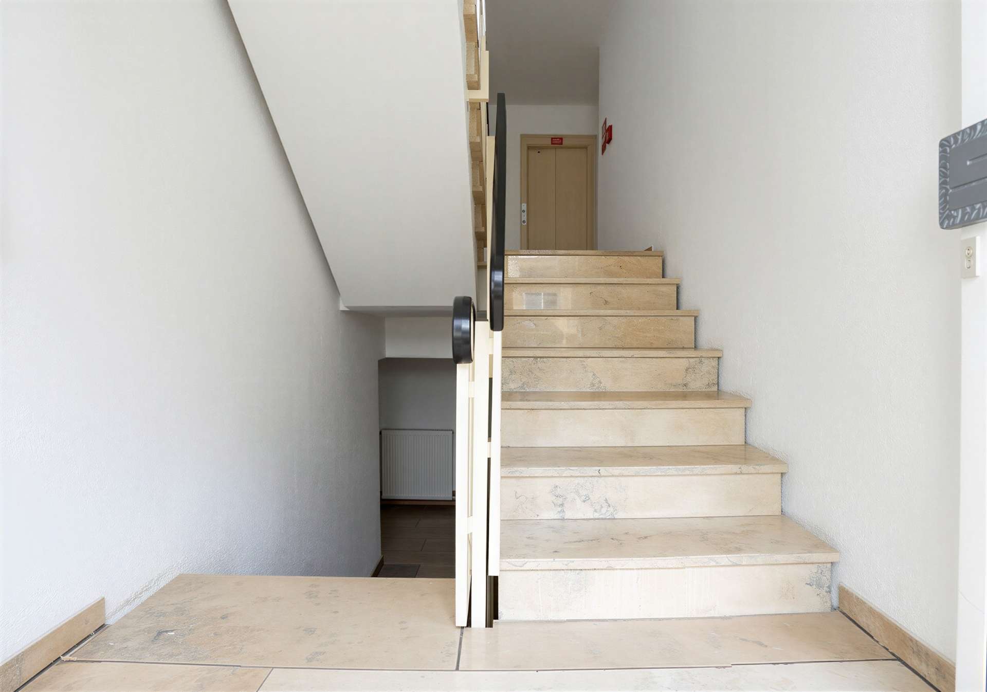 Beige marble staircase in a narrow white-walled hallway, with a black handrail leading up to a wooden door at the top.