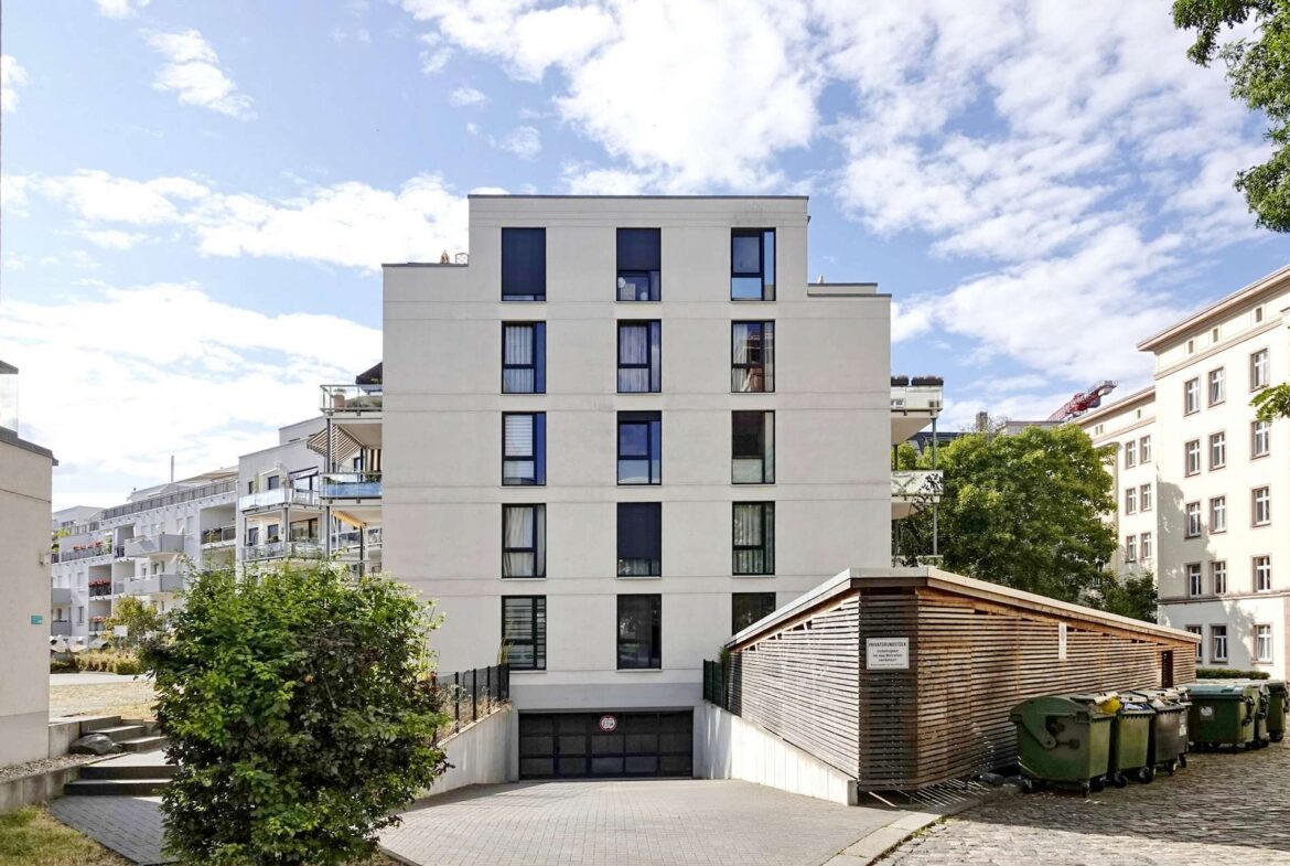 Center-facing white apartment building with multiple windows under a blue sky; a wooden shed and green dumpsters sit to the right, with a cobblestone path in front and surrounding greenery.