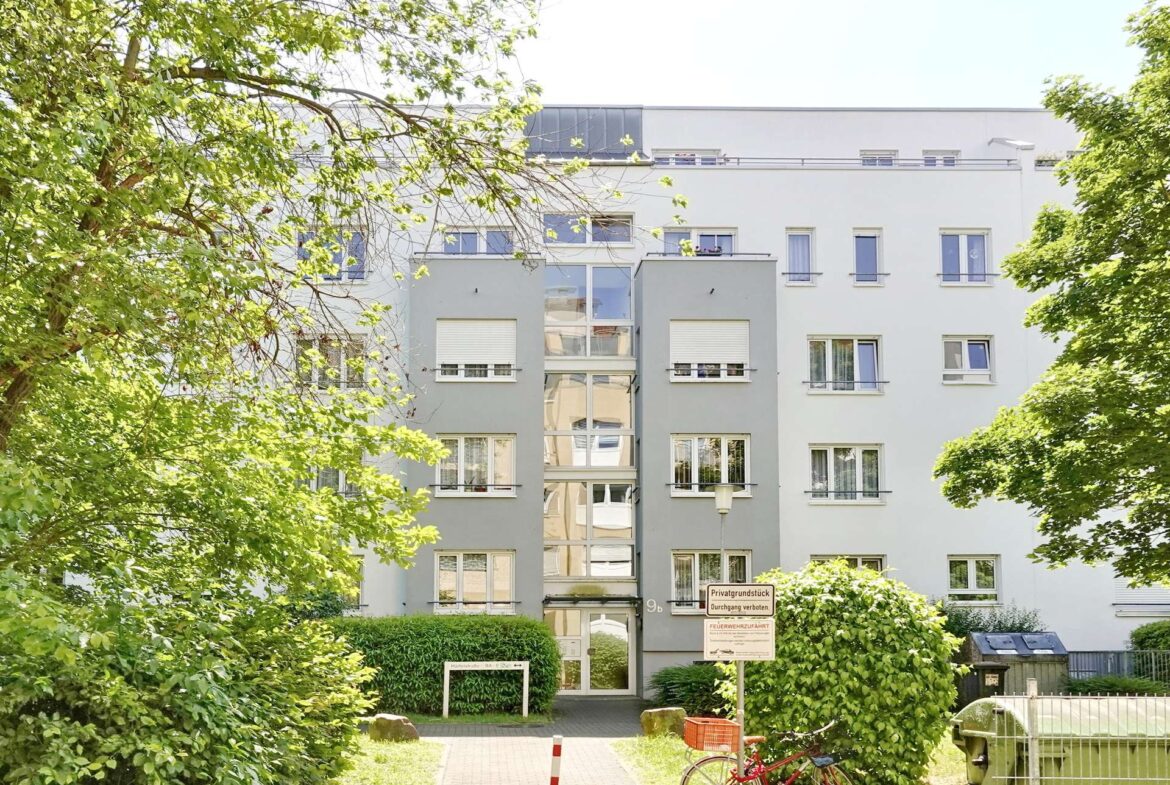 Modern grey apartment building with a central glass stairwell, framed by green trees and shrubbery in a sunny setting.