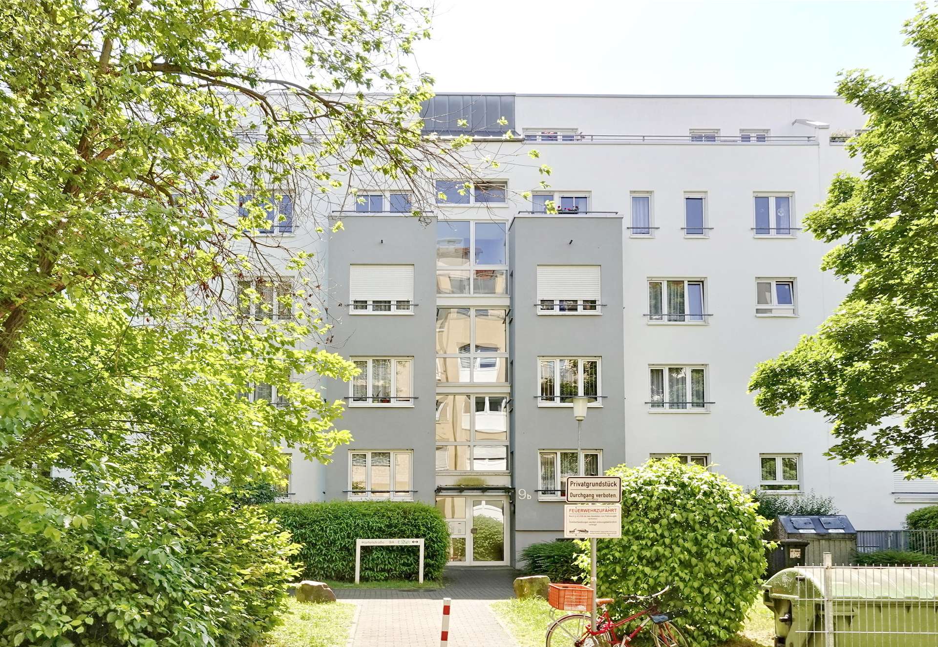 Modern light-grey apartment building with a central glass stairwell, framed by green trees on a sunny day.