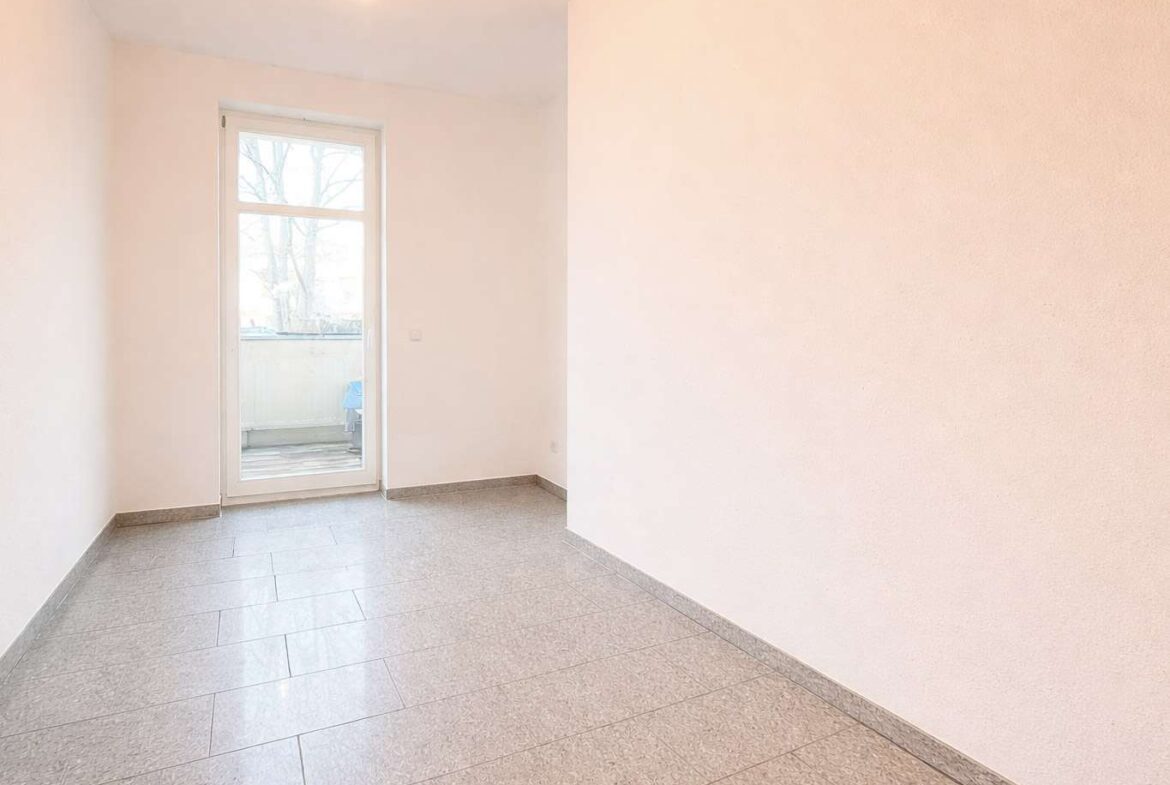 Bright empty room with a glass door to a balcony, pale walls, and gray tiled flooring, lit by a ceiling light.