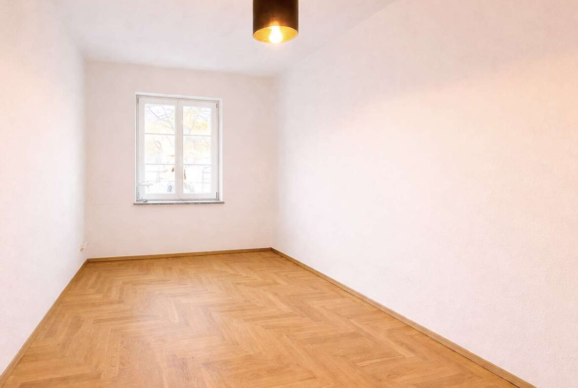 Empty rectangular room with white walls and a light wood parquet floor, a single window on the far wall, and a black pendant light above.