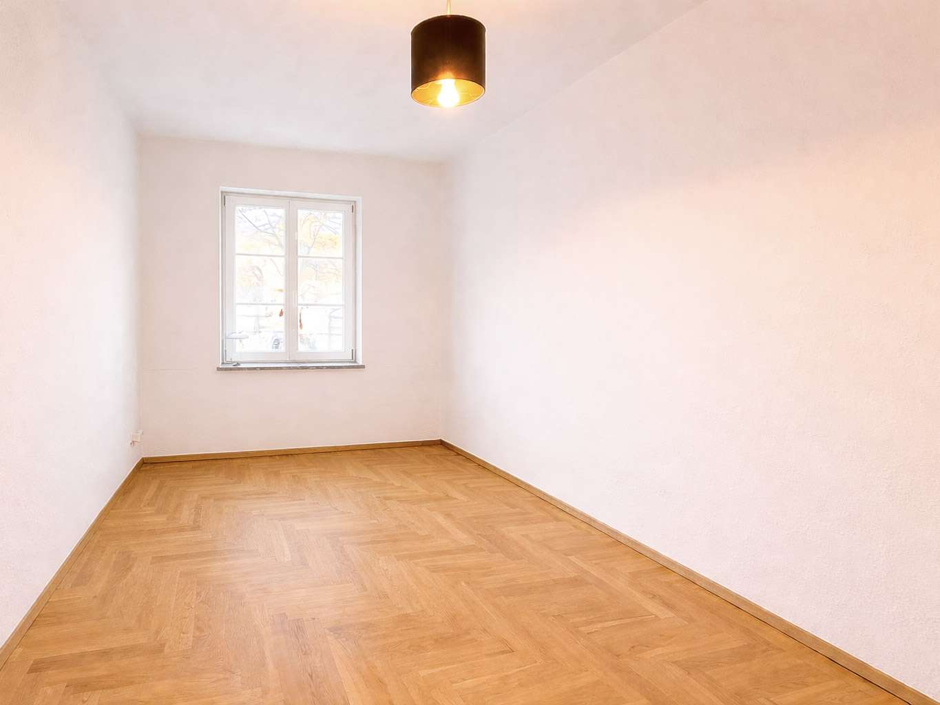Empty rectangular room with white walls and a light wood parquet floor, a single window on the far wall, and a black pendant light above.